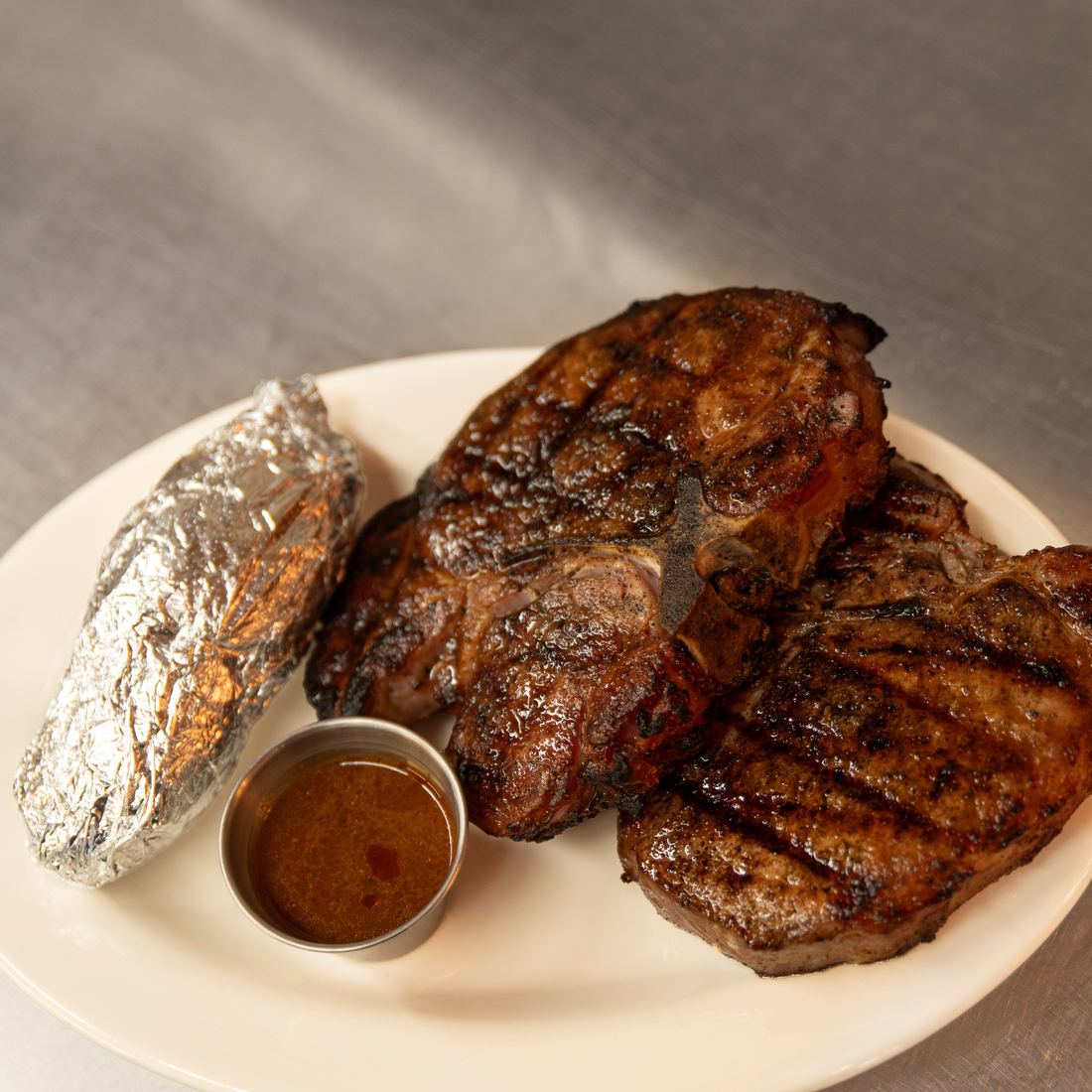Two smoked pork chops with a foil-wrapped baked potato and sauce